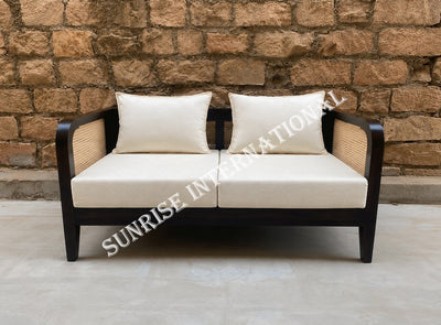 A two-seater outdoor loveseat with cream-colored cushions and a dark brown wooden frame with woven cane side panels, set against a rustic stone wall.