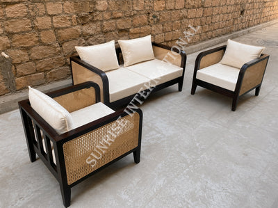 Three-piece outdoor patio conversation set with dark wood frames, woven cane panels, and off-white cushions, arranged on a concrete patio against a stone wall.