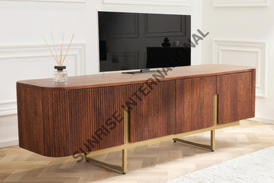 A mid-century modern wooden credenza or TV stand with fluted vertical wood panels and gold metal legs, styled in a bright living room with a television, books, and a reed diffuser.