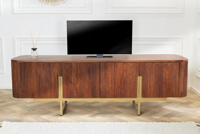 A mid-century modern wooden TV stand with fluted vertical grain and gold metal legs, styled in a living room setting with a television on top.