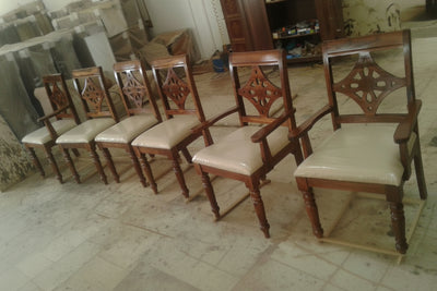 Six wooden dining chairs with cream upholstered seats and carved dark reddish-brown backrests, arranged on a tiled floor in what appears to be a workshop or showroom.